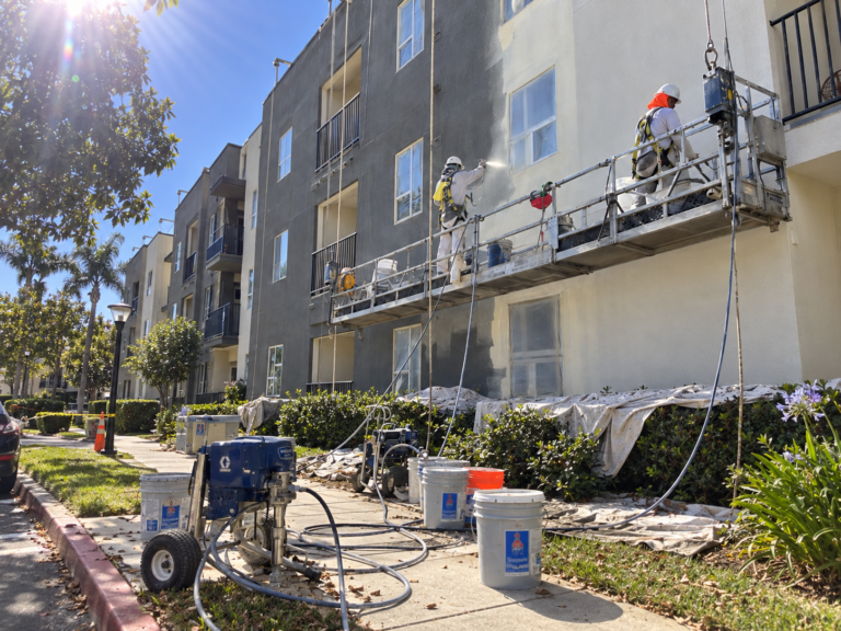 Painters applying an exterior coating to a multifamily building from a suspended work platform.