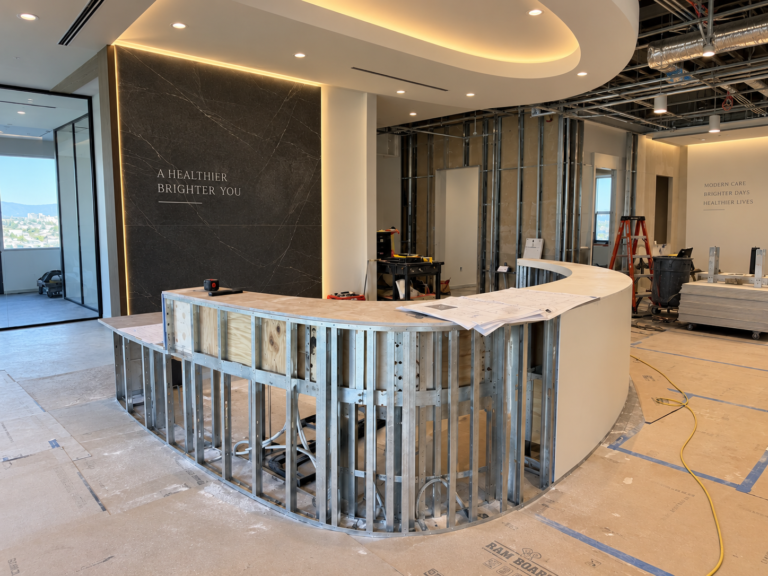 Commercial reception area under renovation with metal framing, exposed ceiling services, and a curved reception desk.